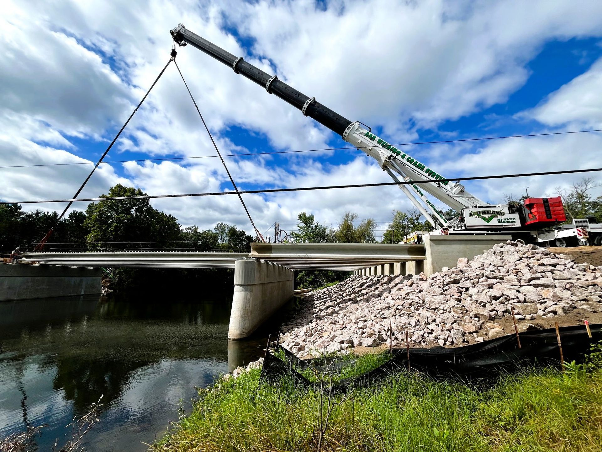 Crane lifting a concrete bridge beam over a river. Rocks line the bank. Blue sky with clouds.