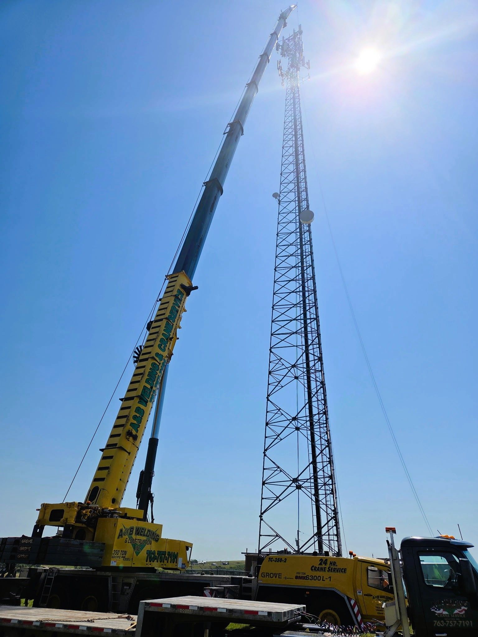 Yellow crane lifting equipment onto a tall communication tower under a bright blue sky.