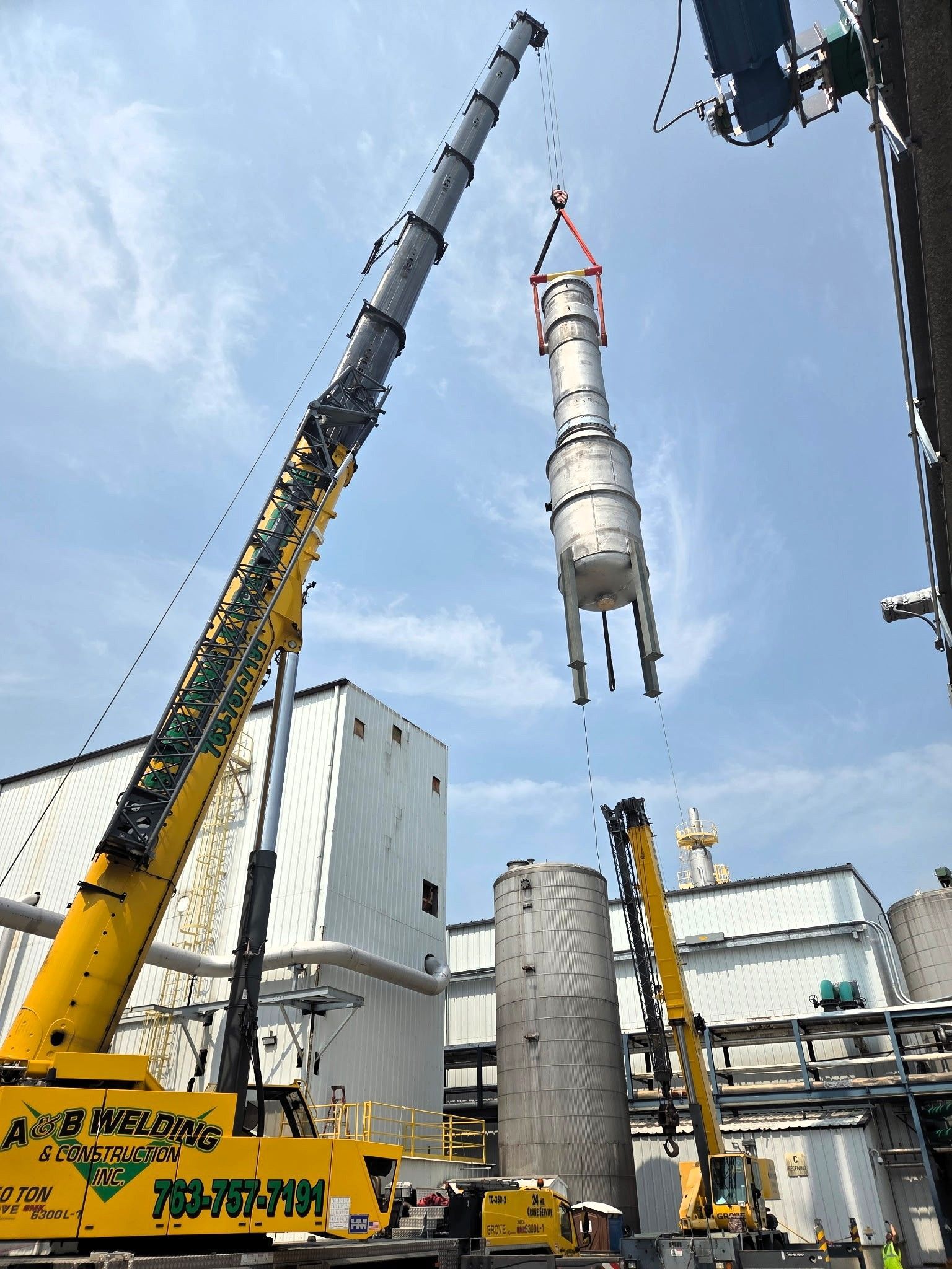 A crane lifts a large cylindrical tank over other industrial structures on a sunny day.
