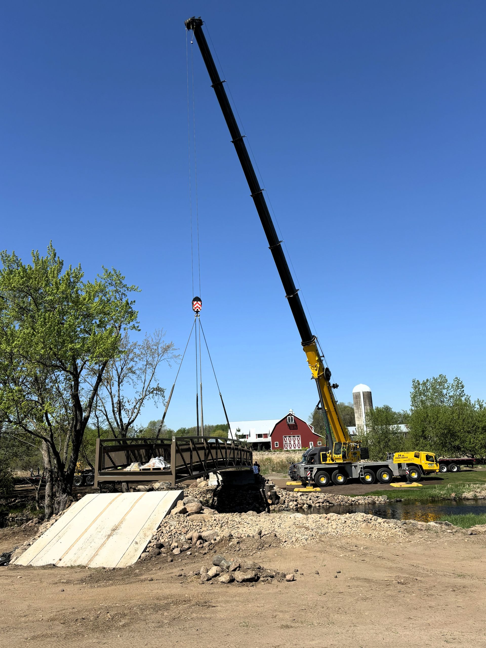 Crane lifting a wooden bridge. Yellow crane against a blue sky, setting with red barn and silo.
