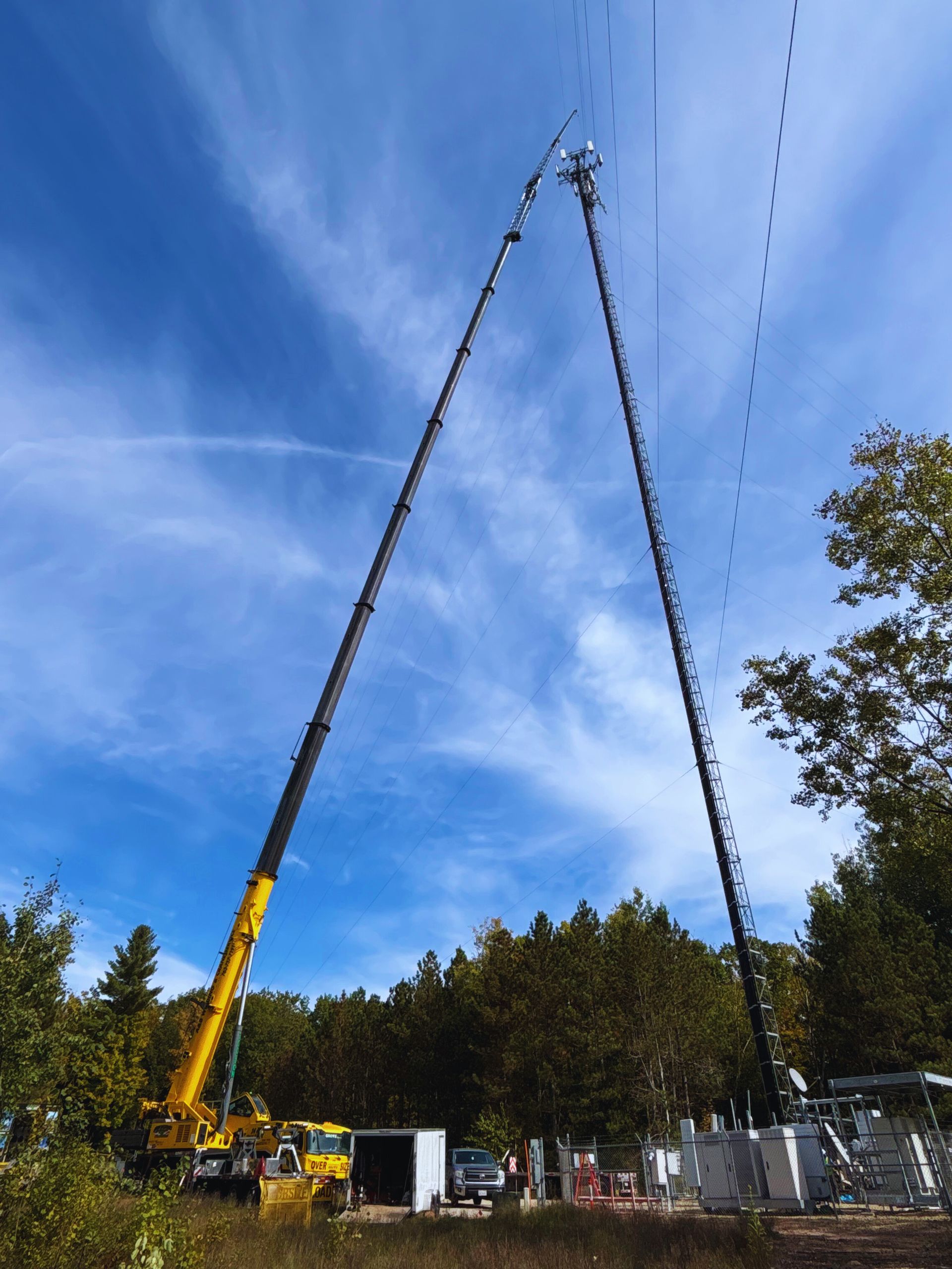 Two cranes erecting a communication tower in a wooded area under a blue sky.
