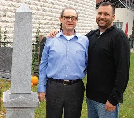 Two men standing beside a headstone. The man on the left has his arm around the other's shoulder.