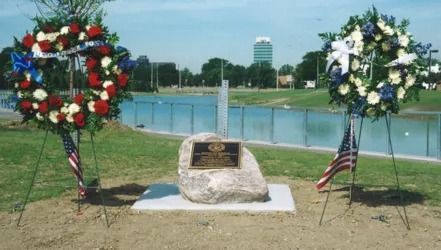 Memorial with wreaths, American flags, and a plaque on a rock, set against a pond and park.