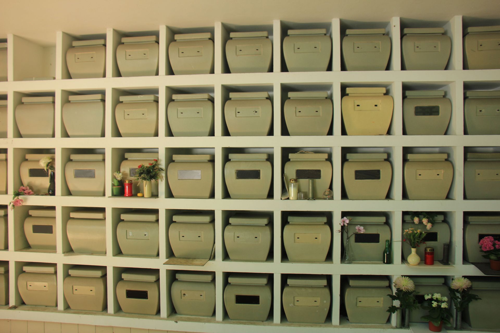 Wall of beige urns in niches, some with floral tributes, within a columbarium.