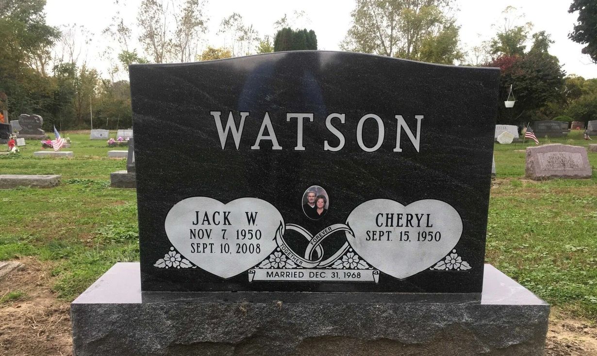 White headstone with flowers, set in a green grassy cemetery with numerous other headstones.