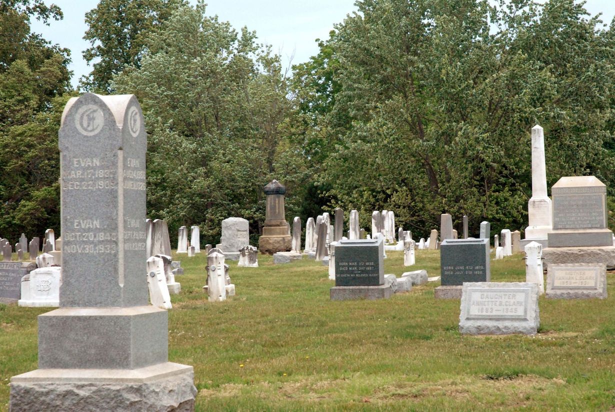 Cemetery with numerous gravestones on a grassy hill, trees in the background, a large double stone marker in the foreground.