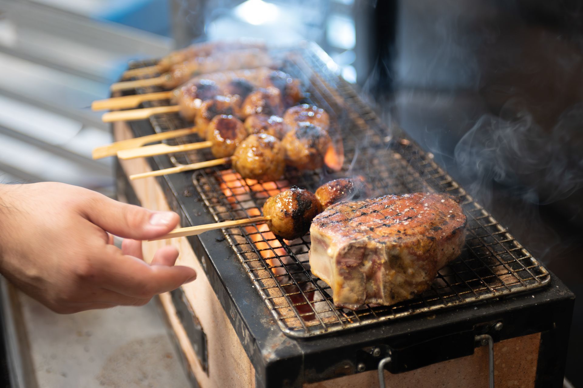 Hand placing skewers and steaks on a smoky grill with charred meat cooking