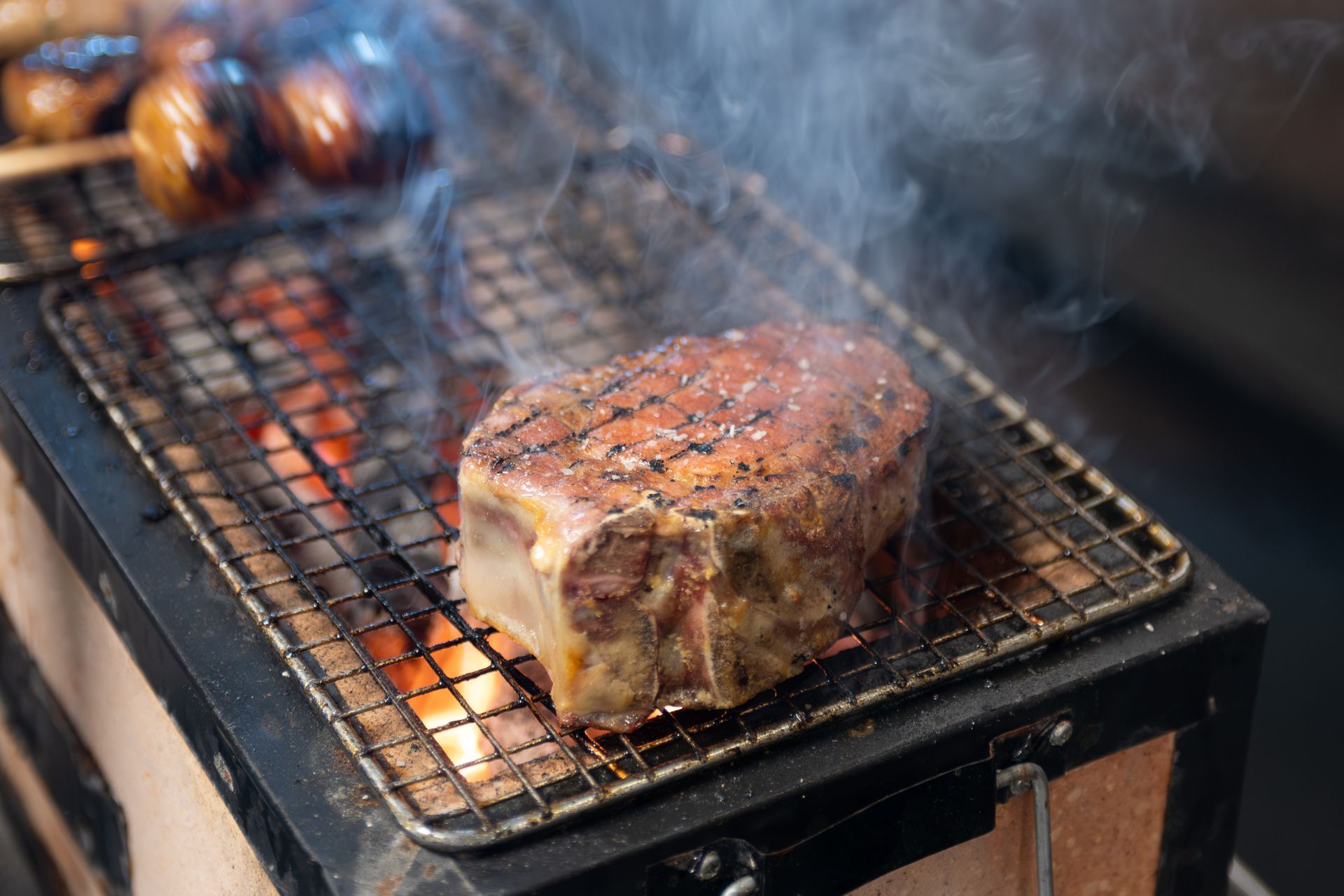 Steak grilling on a wire rack over open flames, with smoke rising and roasted vegetables in the background