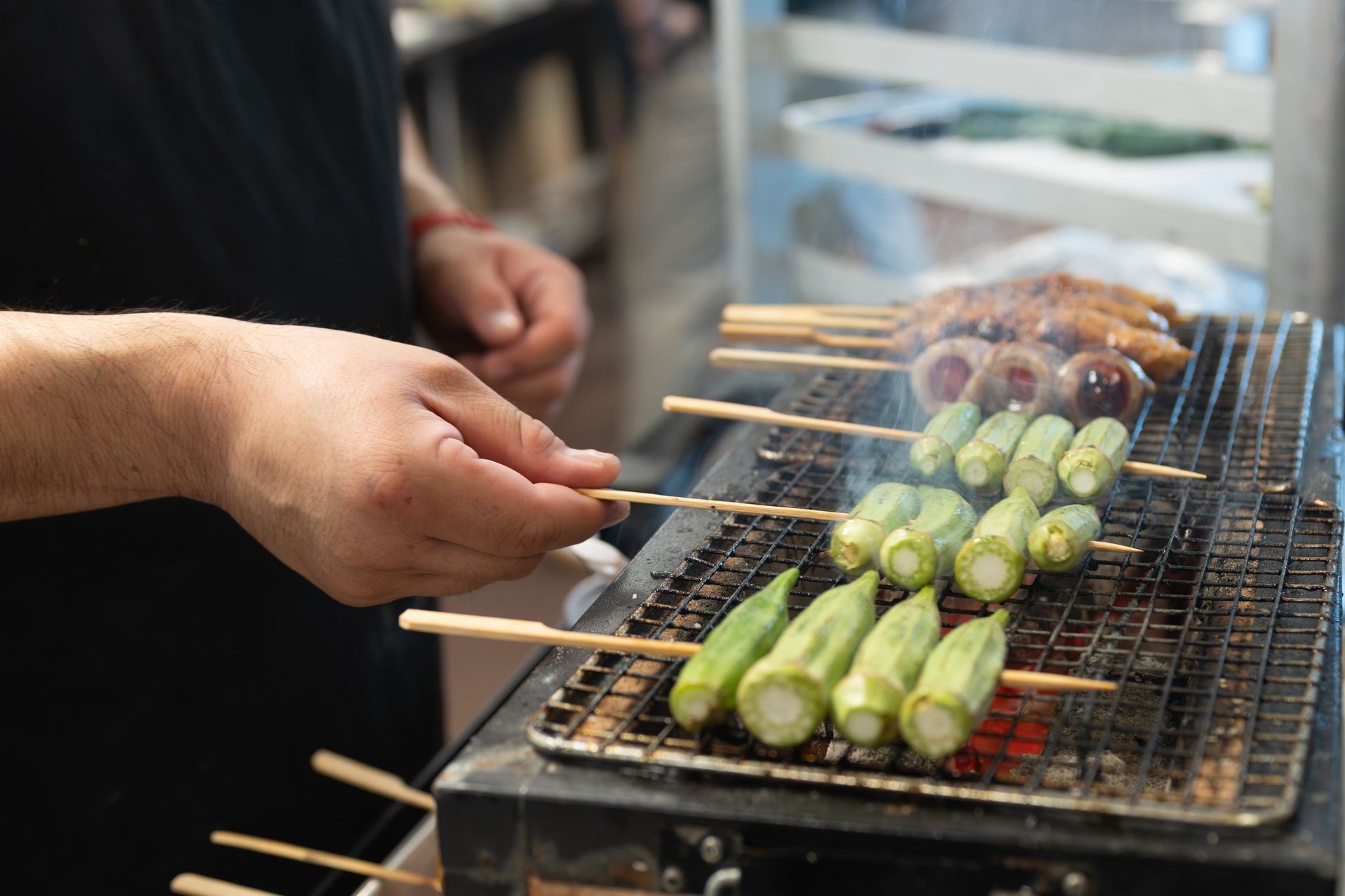 Hands grilling skewered zucchini over a charcoal barbecue with visible smoke