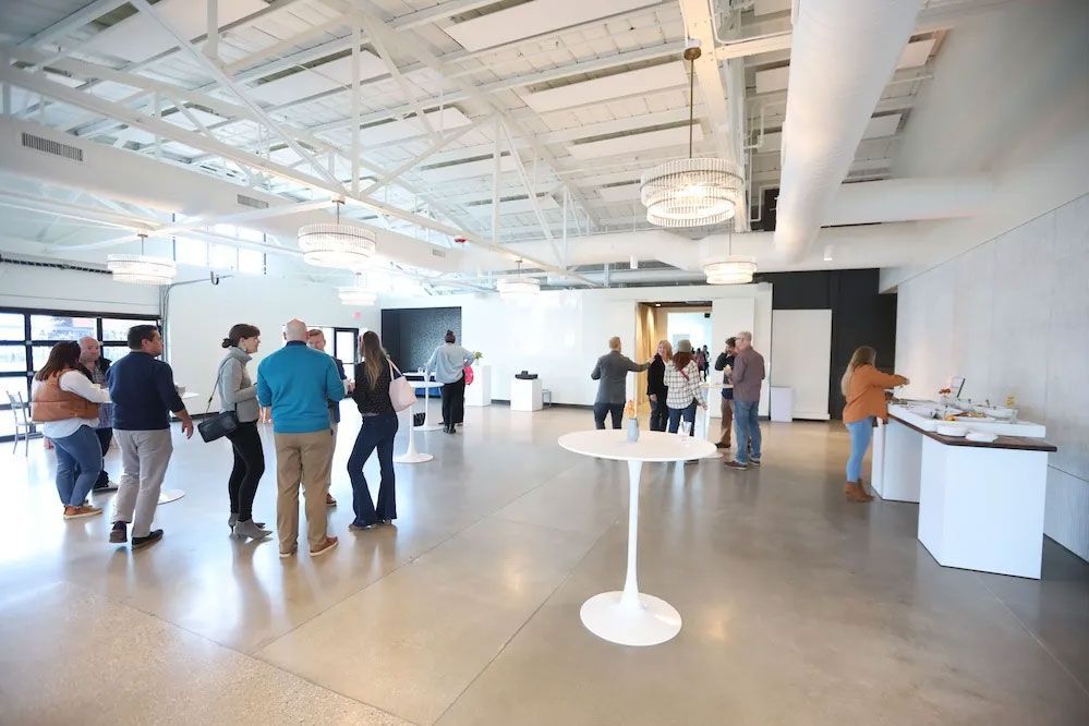 People gathering in a large, modern event space with white walls, polished concrete floor, and round tables.