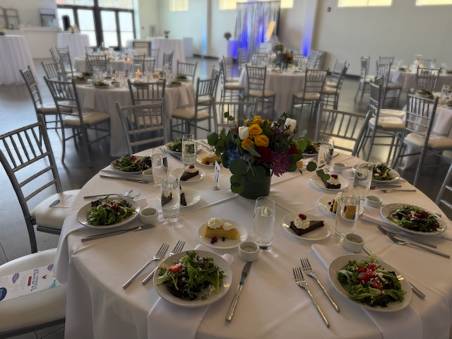 Round tables set for a formal event, with salad, dessert, and floral centerpiece. Silver chairs.