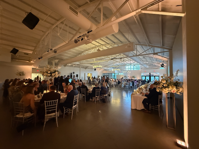 Wedding reception in a large white venue. Guests seated at round tables, white flowers, overhead lighting.