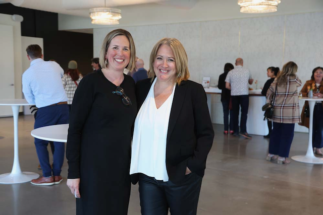 Two women smile, posing together at an event with others in the background.