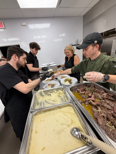 Chefs plating food from serving trays in a commercial kitchen. One person is passing food to another, while others serve.