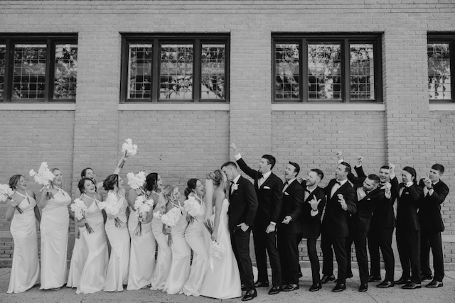 Wedding party celebrates outside a brick building. Bride and groom kiss, bridesmaids hold bouquets, groomsmen cheer.