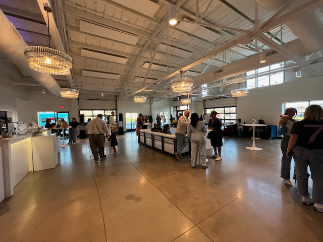 Large event space with people mingling. Chandeliers hang from a white ceiling, open garage doors, and a bar.