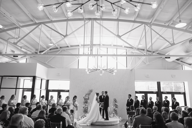 Wedding ceremony in a modern hall; bride and groom at altar, guests seated, high ceiling with lights.