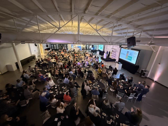 Large event in a white-walled venue; many people seated at tables, a stage with a presentation.