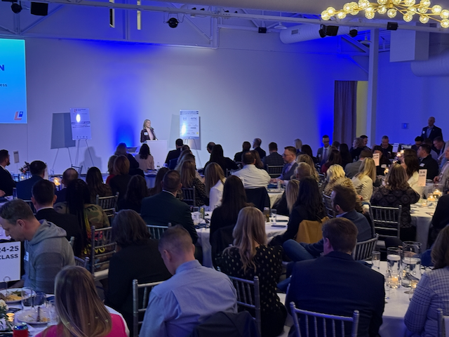 A presentation in a large hall with people seated at round tables. A woman speaks at the front.