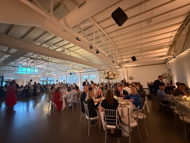 Large event hall with tables, people dining. Overhead lighting, floral centerpieces.
