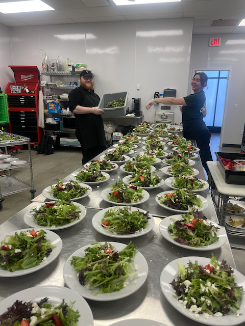 Two people preparing many salads on a long stainless steel table in a bright kitchen.