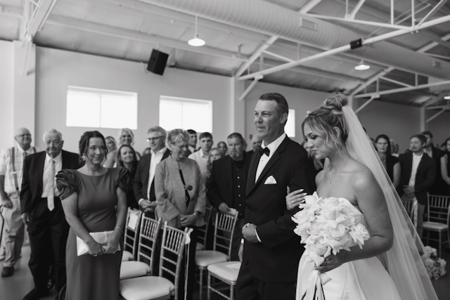Bride escorted by her father down the aisle at a wedding ceremony, guests watching in an indoor venue.