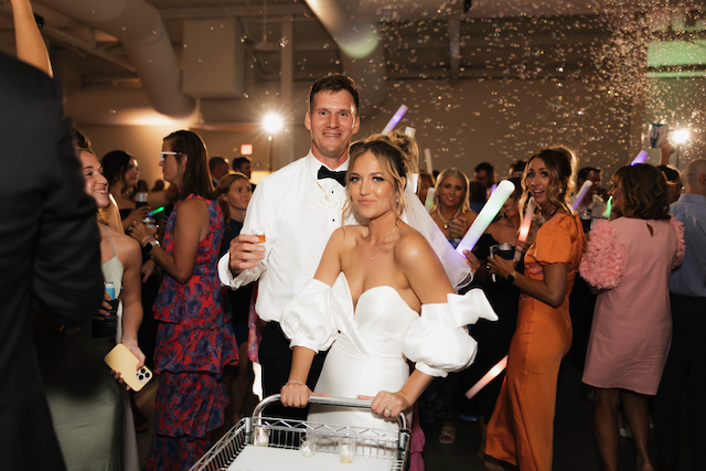 Bride and groom pose with shopping cart at wedding reception, surrounded by guests holding glow sticks and confetti.