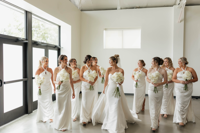 Bridesmaids in white halter dresses with bouquets walk in a bright room with large windows.