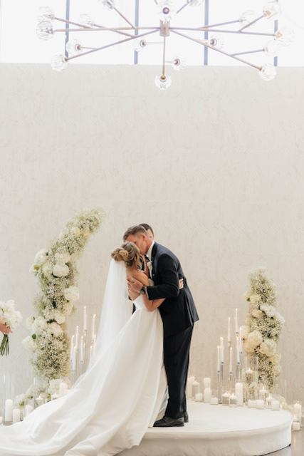 Couple kissing at a wedding ceremony; bride in white dress and veil, groom in black suit, floral backdrop.