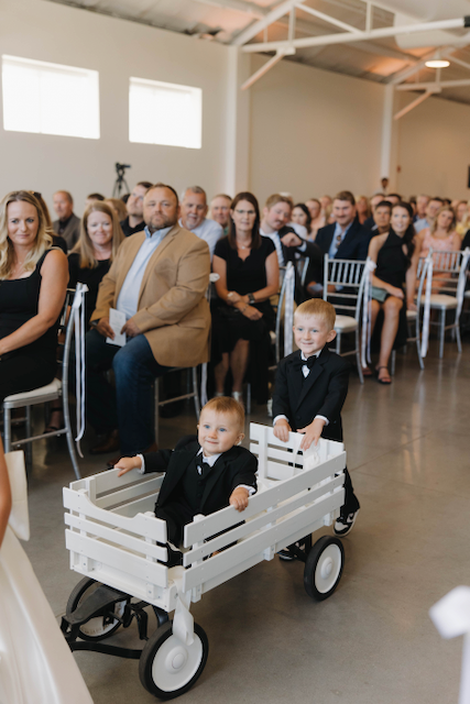 Two boys in a white wagon at a wedding; guests in the background.