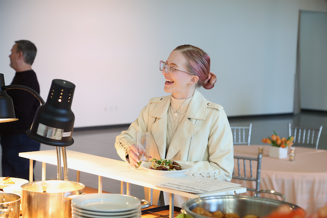 Woman laughs while holding a plate of food at a buffet. She wears glasses and a tan jacket.