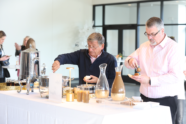 Two men serving themselves coffee from a buffet table; light-colored room with windows.