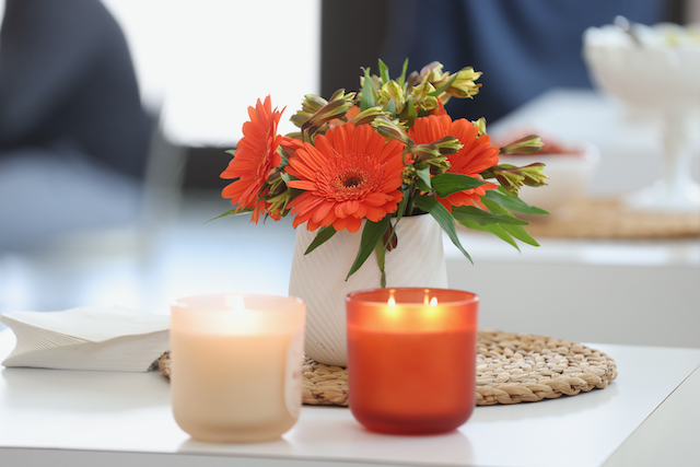 Close-up: orange flowers in white vase, lit candles on a white table.