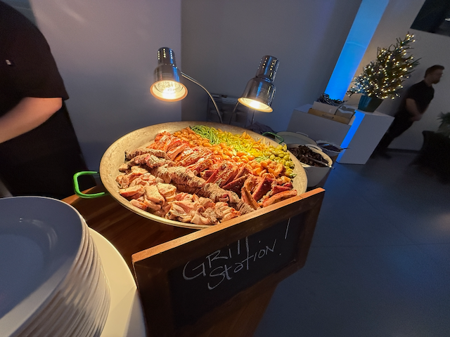 Grill station with meat and vegetables, under warming lights. People are nearby.