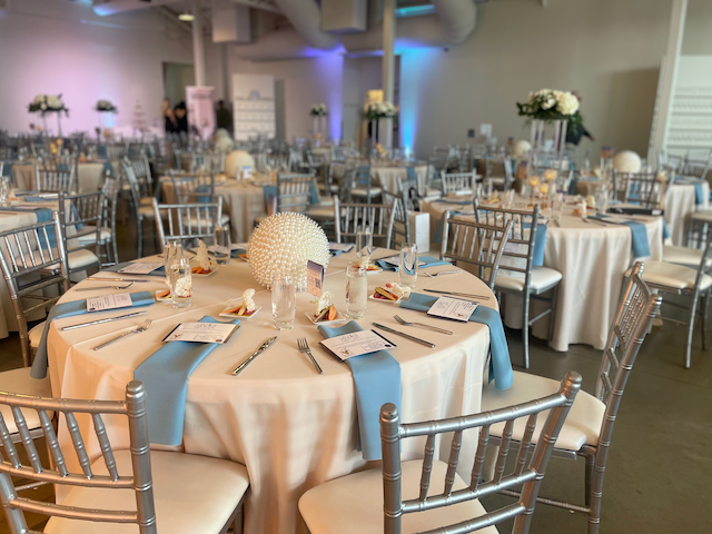 Elegant event hall with round tables set for a celebration. Tables have cream linens, blue accents, and silver chairs.