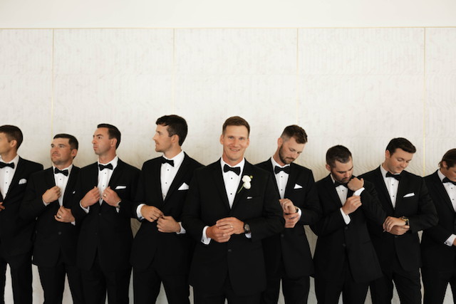 Men in tuxedos posing in front of a white wall, adjusting their jackets.