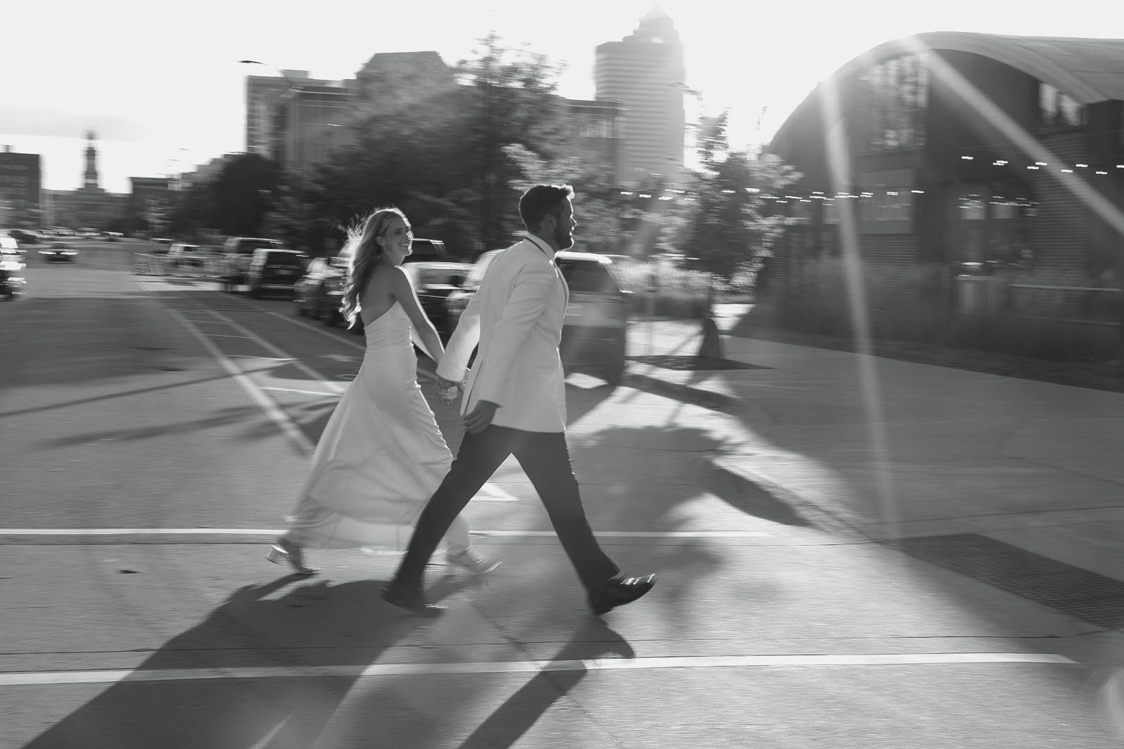 Two people crossing a sunlit street, one in a light dress and one in a suit, with long shadows on the pavement.