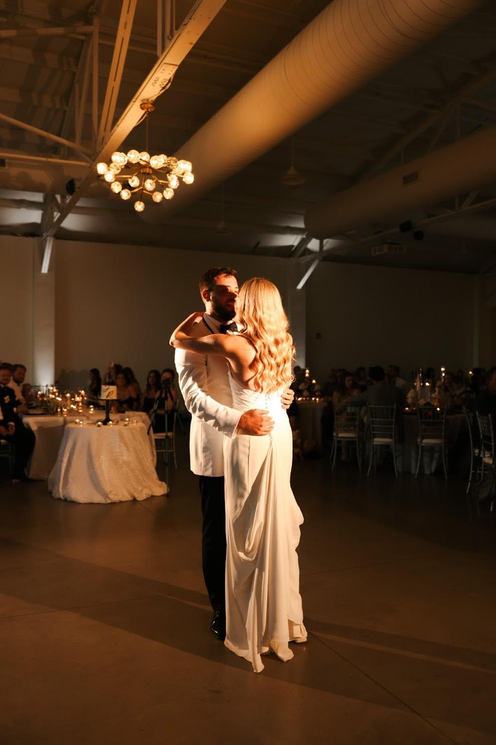 Couple slow dancing at a dimly lit wedding reception, with warm chandelier light overhead.