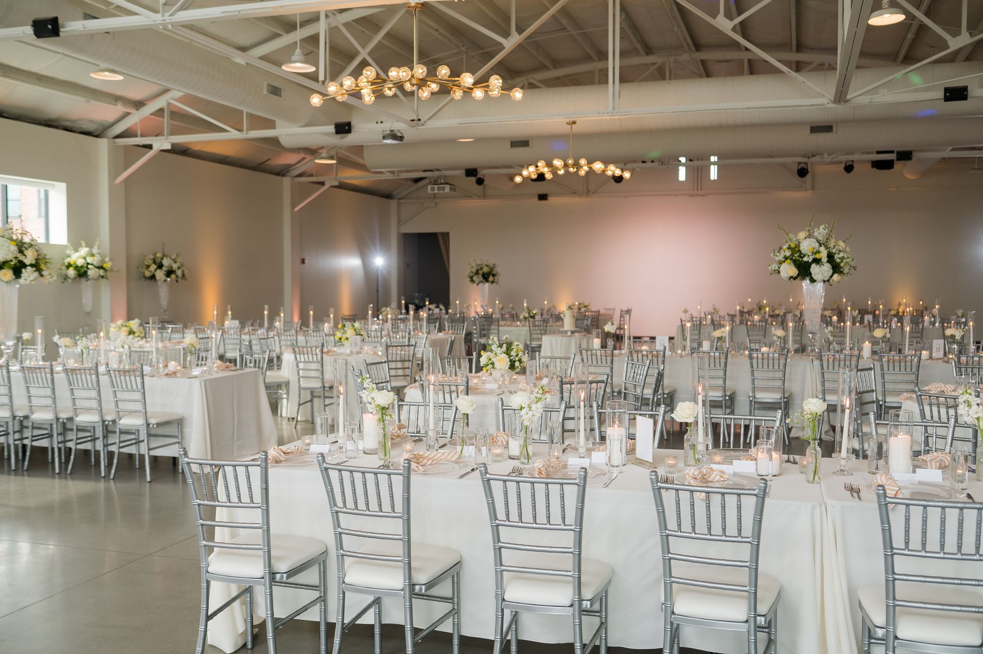 Elegant wedding reception hall with round tables, white chairs, floral centerpieces, and chandeliers.