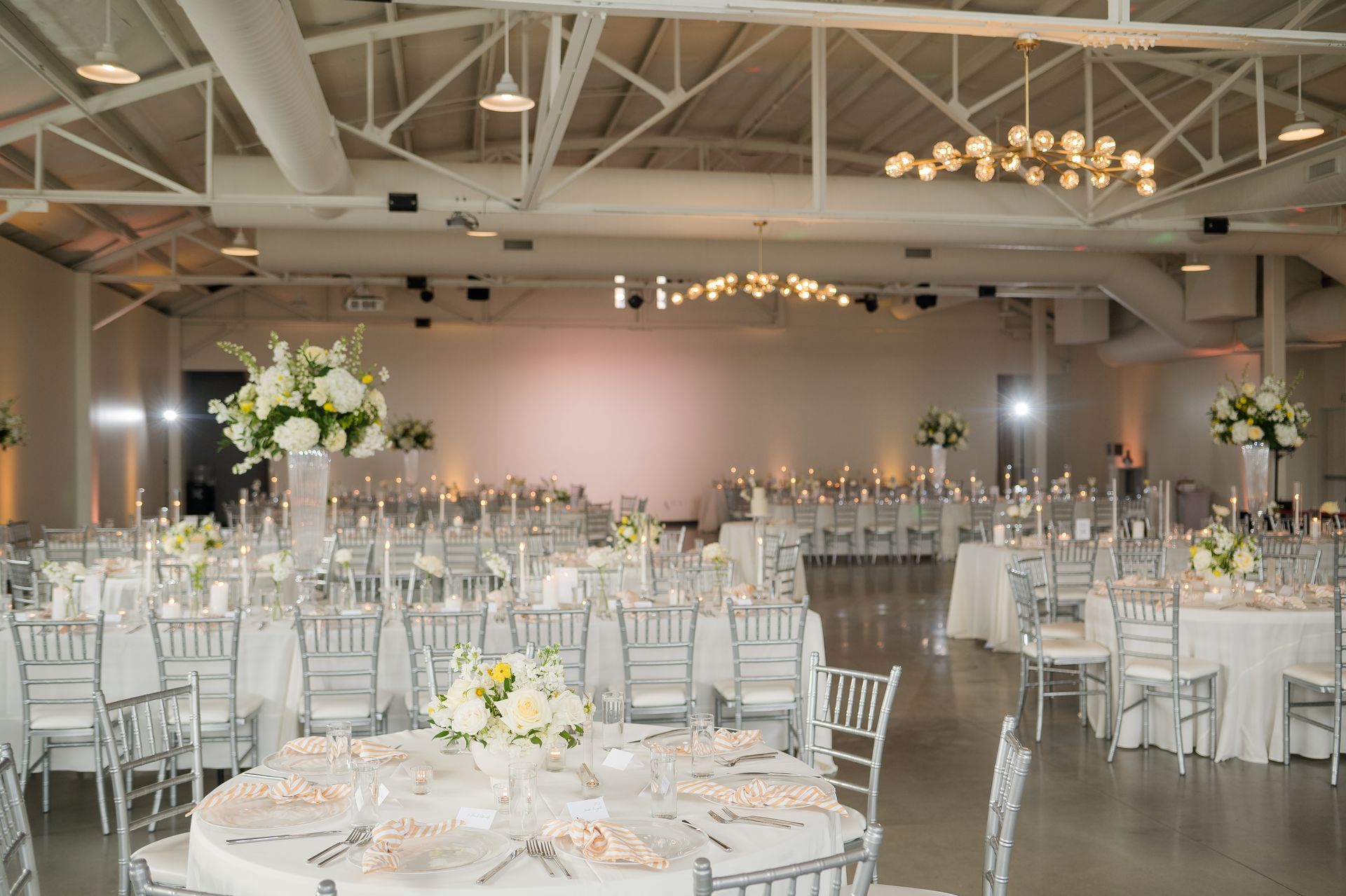 Elegant reception hall with round tables, white chairs, floral centerpieces, and soft chandelier lighting.