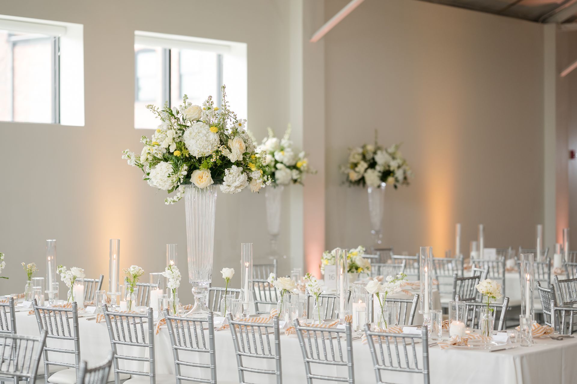 Elegant wedding reception with white floral centerpieces, clear chairs, and candlelit tables in a bright ballroom.