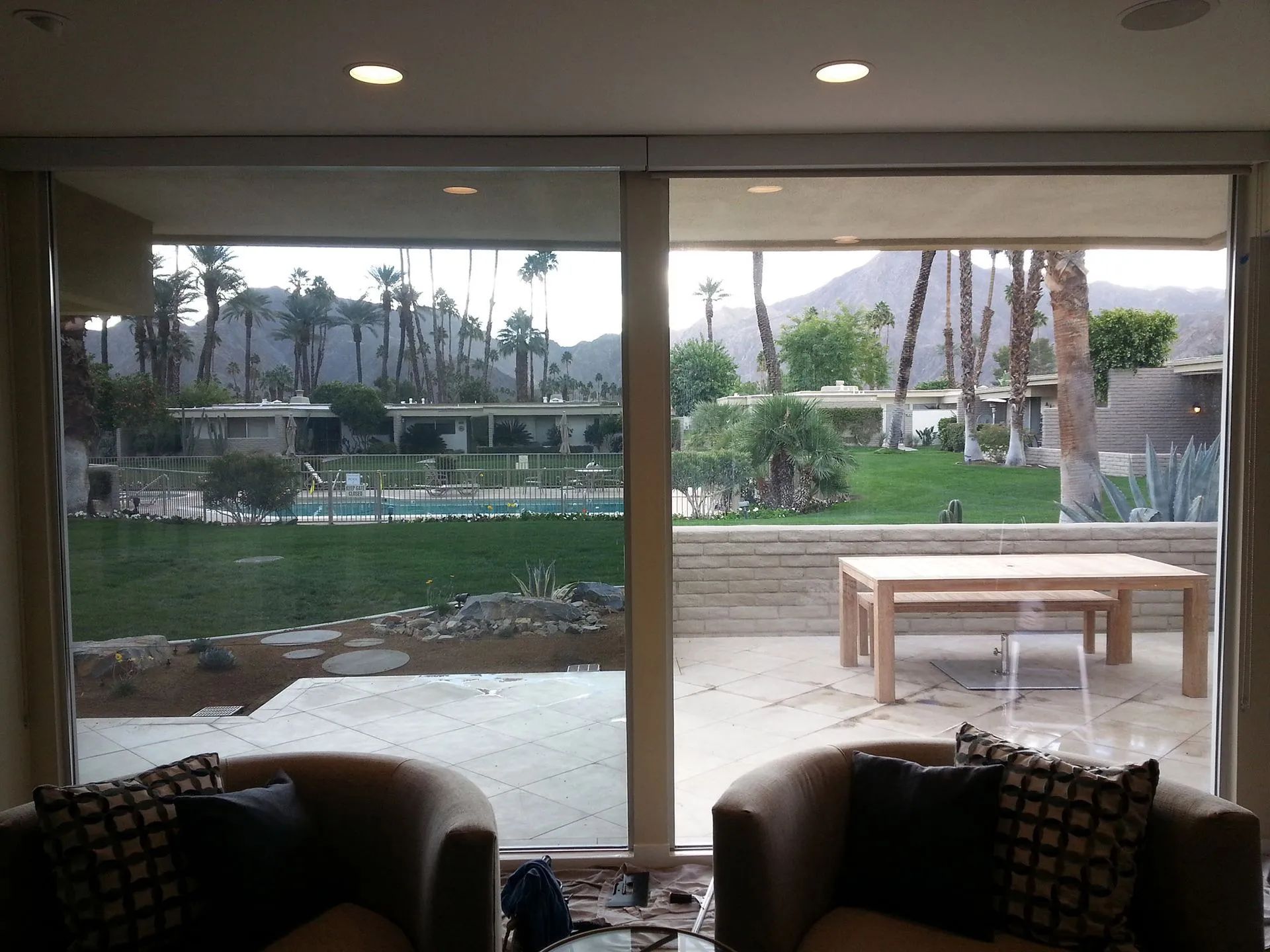 View through sliding glass doors, of backyard with pool, patio table, and mountain backdrop. Interior, two chairs.