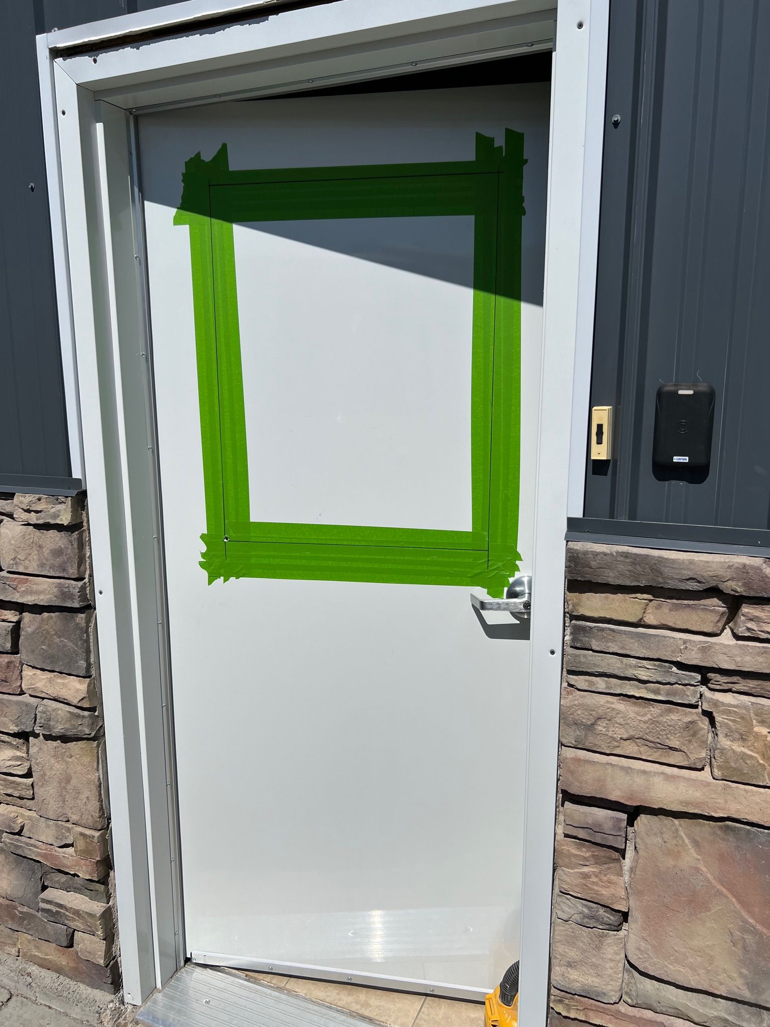 White door with green tape square, set in a gray frame, mounted to a stone exterior wall.