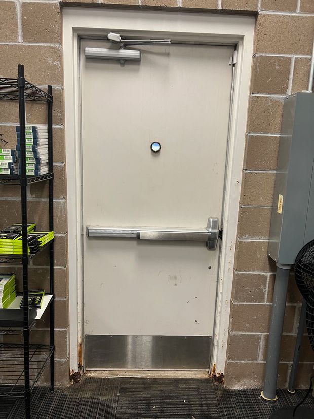 Metal door with push bar, emergency exit sign, and a metal kick plate set in a brick wall.