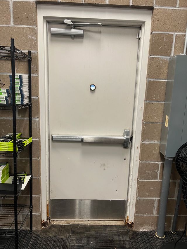 Metal door with push bar, emergency exit sign, and a metal kick plate set in a brick wall.