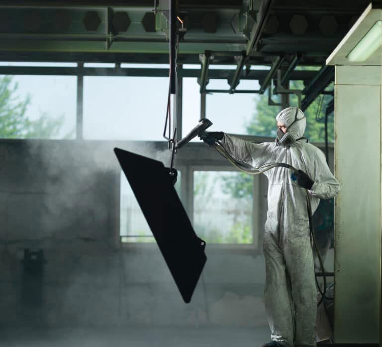 Person in protective suit spray painting a black object in a factory.