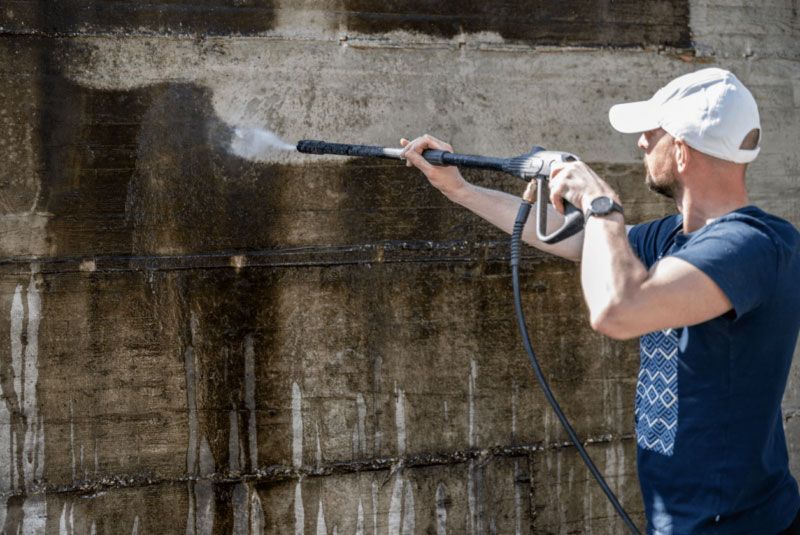 Man pressure washing a dirty concrete wall with a white hat and blue shirt.