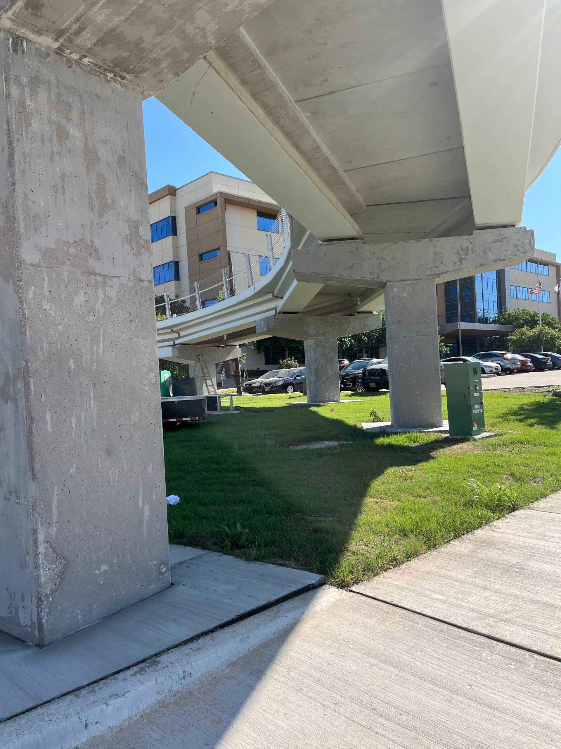 Concrete supports for an elevated train track, green grass, sidewalk, sunny day.