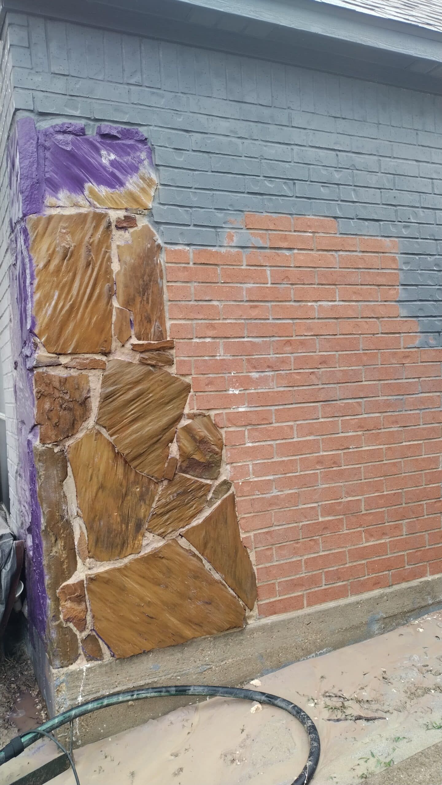 Exterior wall with brick, stone, and painted sections; a water hose lies in front.