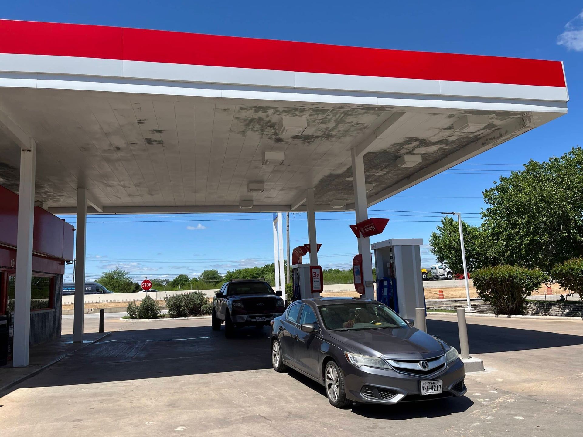Cars at a gas station, under a red and white canopy on a sunny day.